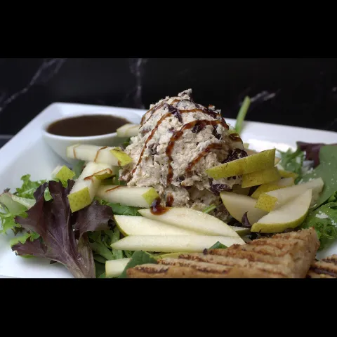 A scoop of chicken salad with dried cranberries drizzled with balsamic glaze, served over mixed greens with fresh sliced pears, grilled flatbread triangles, and a ramekin of dressing on a white plate.