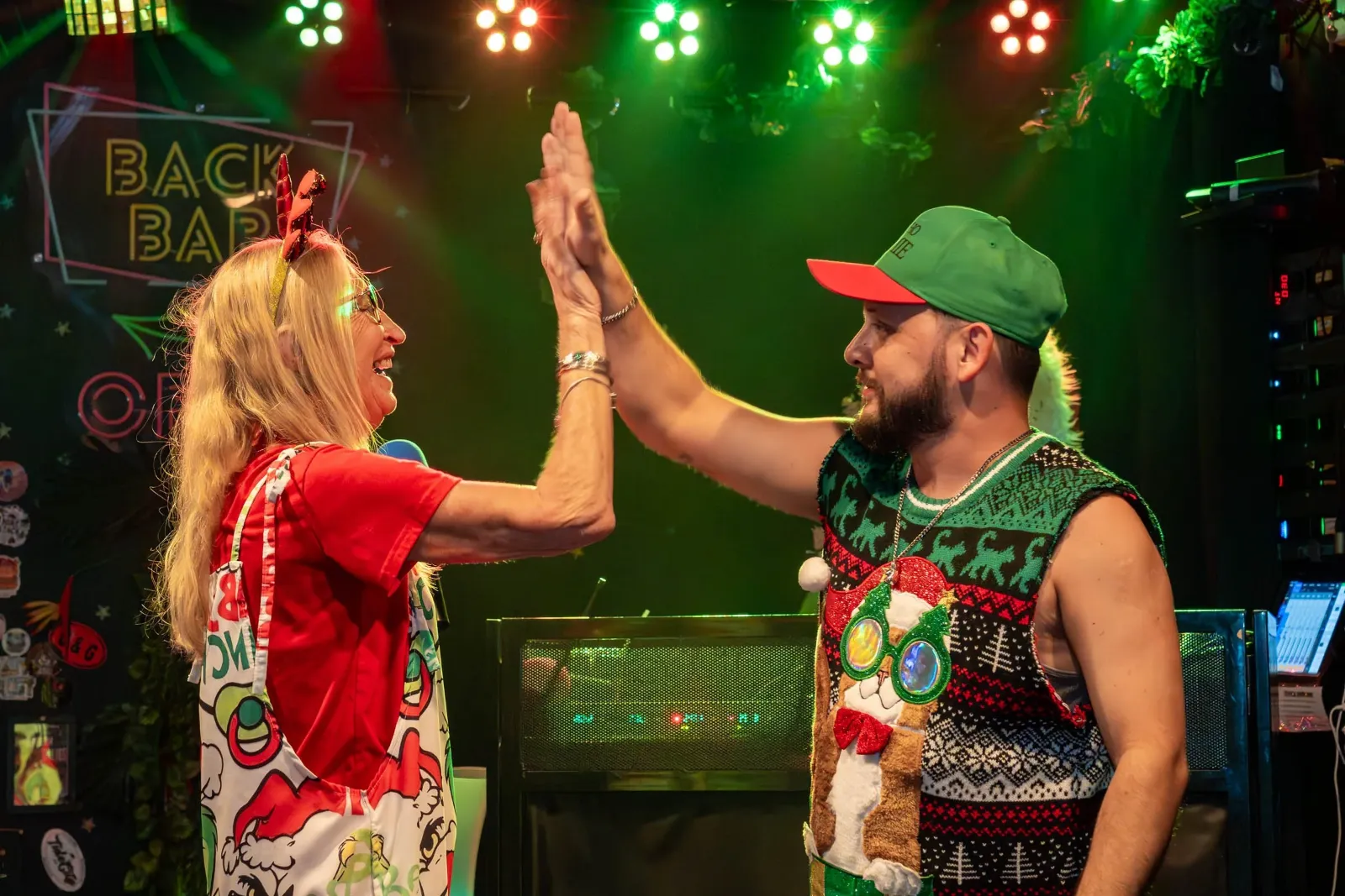 A woman and a man high-fiving each other on a stage decorated with Christmas and holiday themed items, colorful lights overhead, and a sign that says 'Back Bar' in the background.