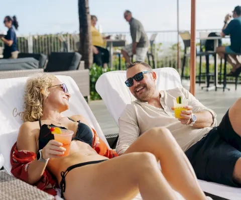 A couple is relaxing on loungers by a pool, smiling and holding cocktails.