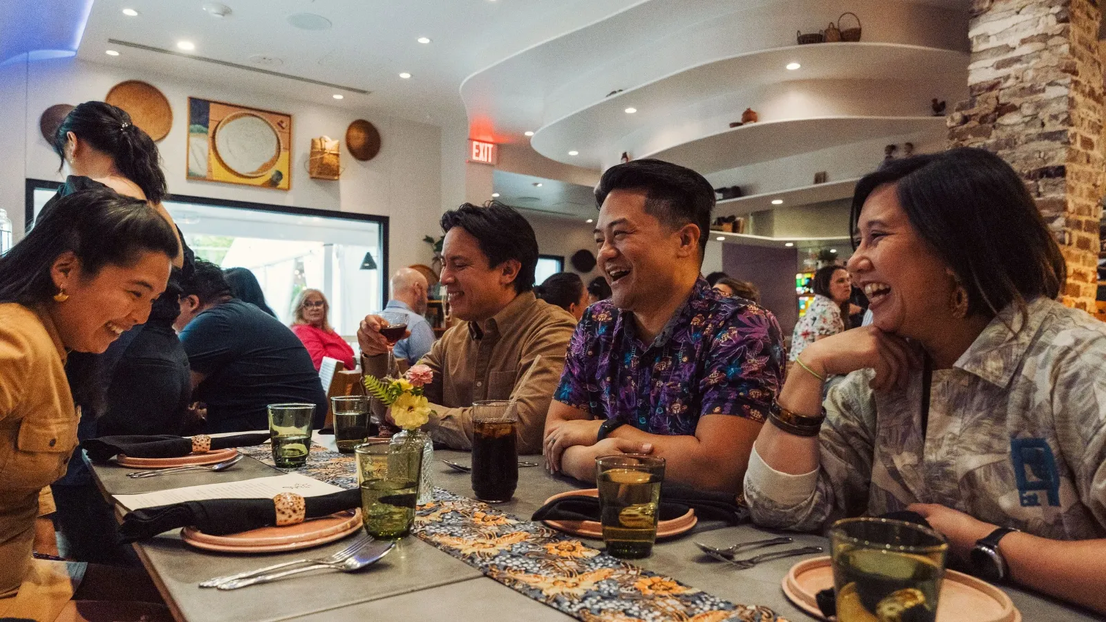 A group of people sitting at a dining table in a restaurant, smiling and laughing together. The table is set with plates, glasses, and cutlery, with a decorative runner and flowers. The restaurant has