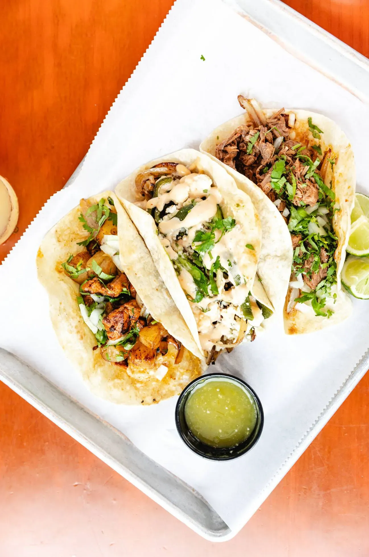 Three tacos are sitting on a white plate on a wooden table.