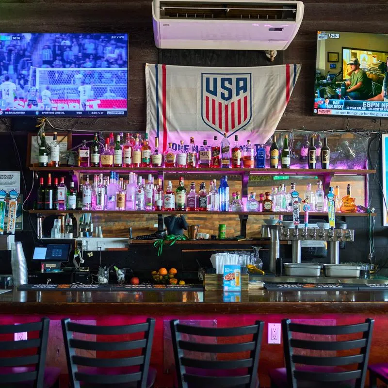 Bar area featuring a wooden counter, dark slatted bar stools and a lit liquor display shelf.