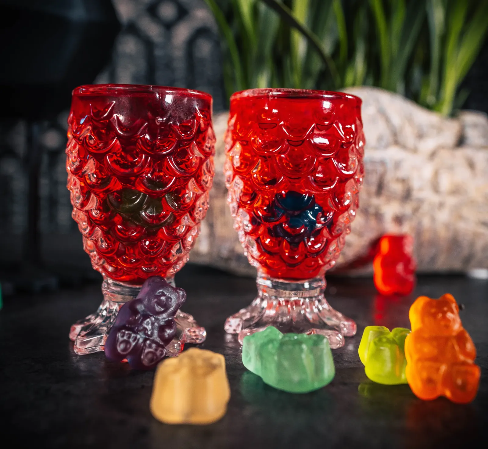 Two red glass goblets filled with gummy bears, surrounded by colorful gummy bears on a dark surface.