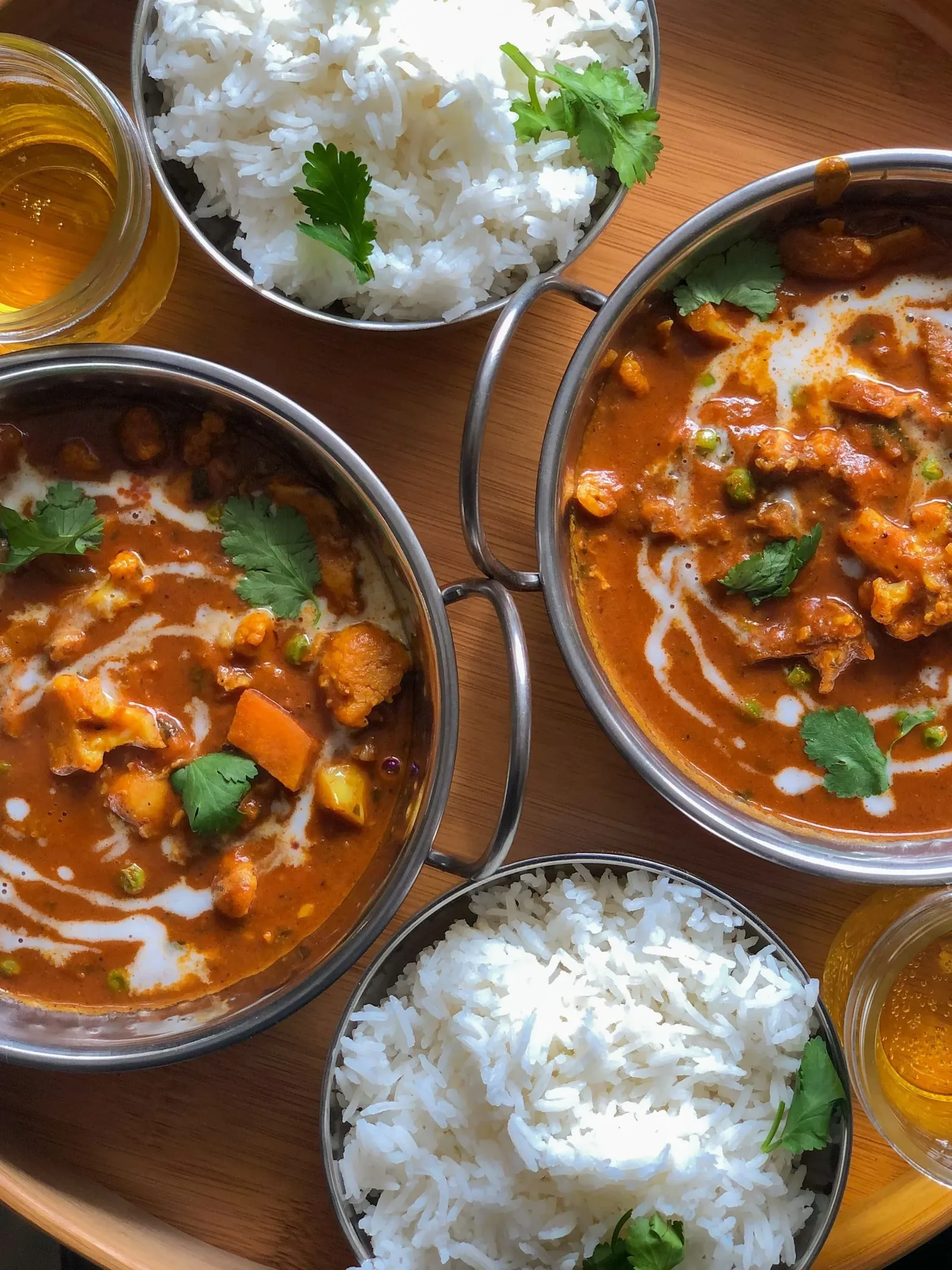 Two bowls of curry with vegetables in a rich orange sauce, garnished with cilantro, alongside bowls of white rice and two glasses of a yellow drink on a wooden tray.