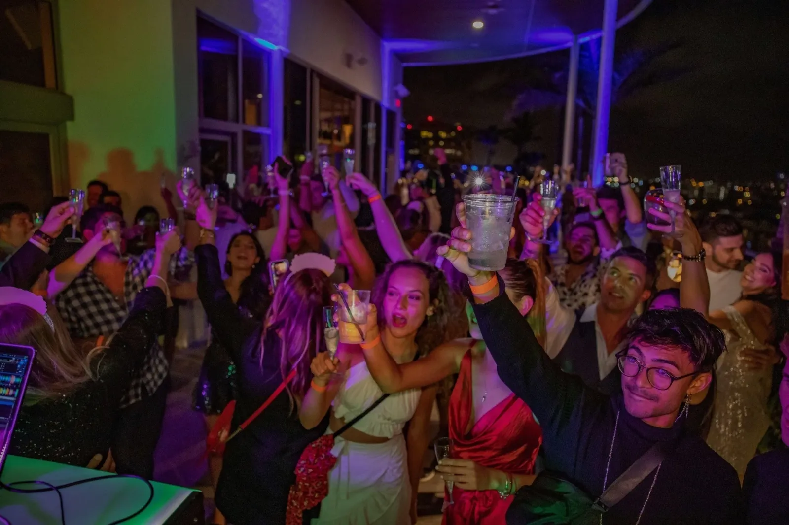 People partying at a nightclub, raising glasses in celebration with colorful lighting and a view of city lights outside.