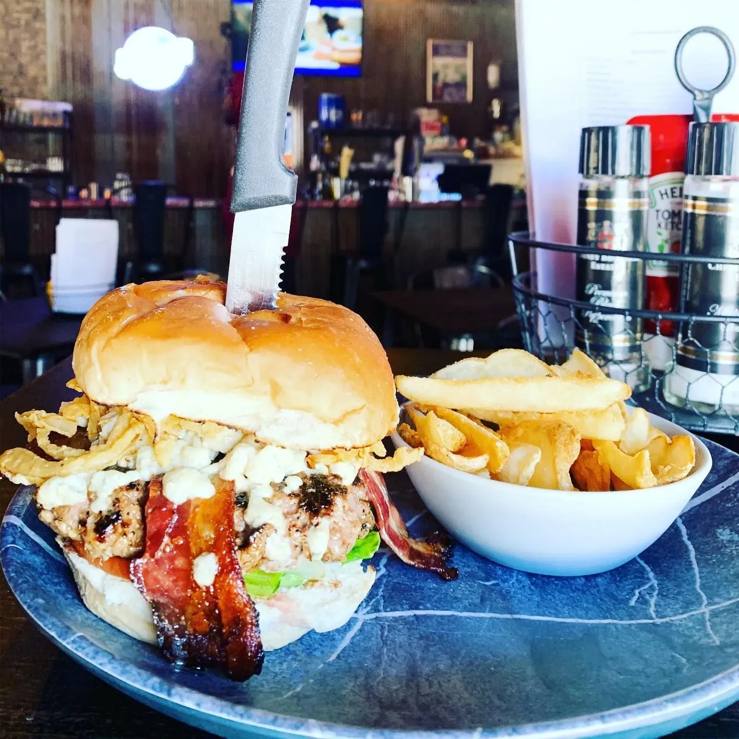 A burger with bacon and onion strings, skewered with a knife, next to a bowl of french fries on a blue marbled plate.