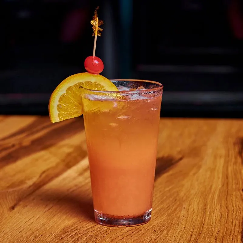 An orange cocktail drink glass garnished with an orange wedge and cherry.