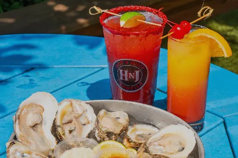 Two colorful cocktails, one red and one orange, with garnishes of lime, cherries, and orange slices, alongside a plate of raw oysters with lemon slices on a blue table.