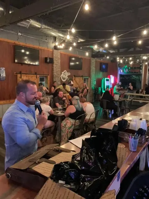 A person in a light blue shirt speaks into a microphone at an event inside a rustic, dimly lit bar with string lights.