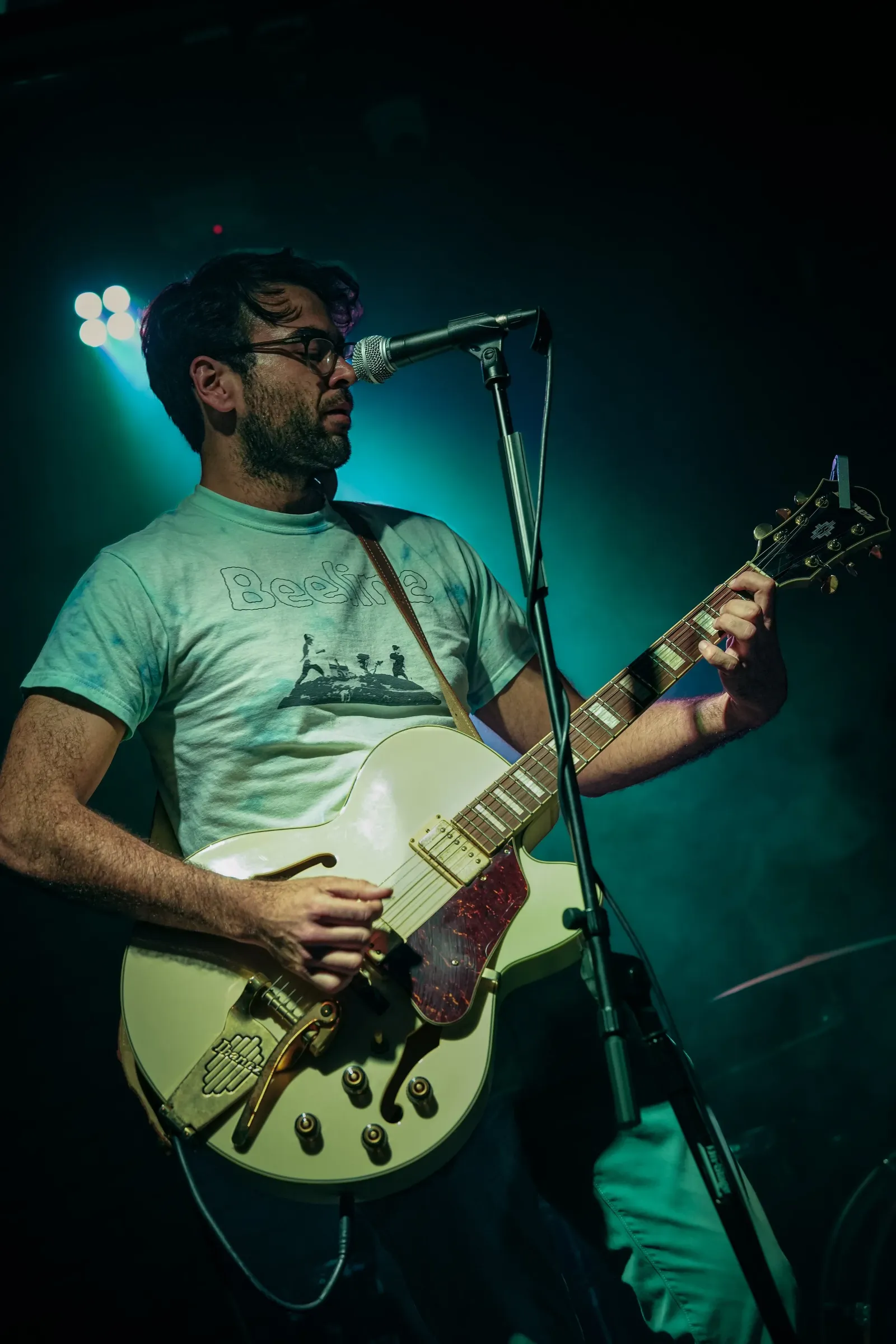 A man with dark hair, beard, and glasses playing an electric guitar on stage with green and blue lighting.