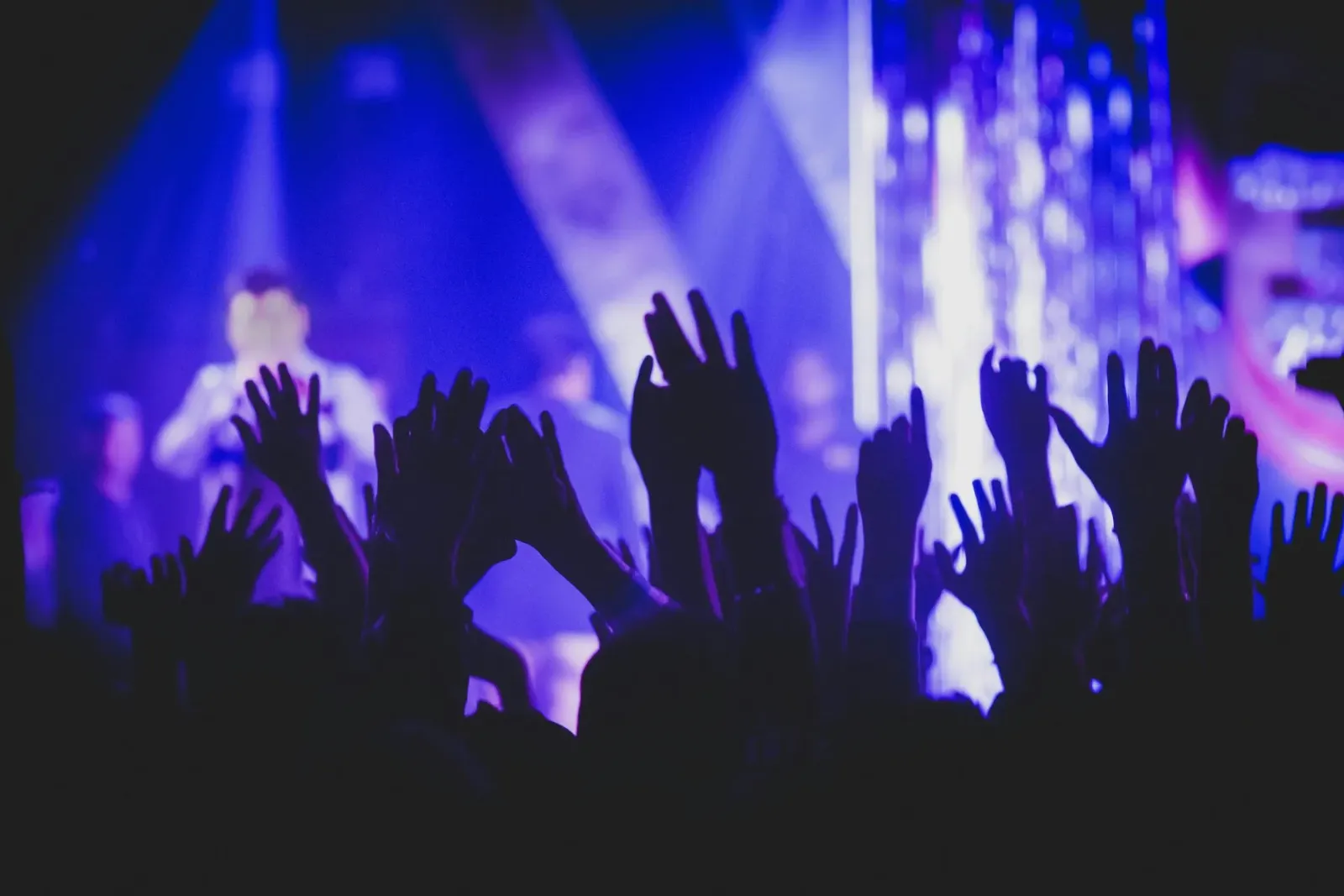 Silhouetted hands raised towards a stage bathed in blue light at a concert.