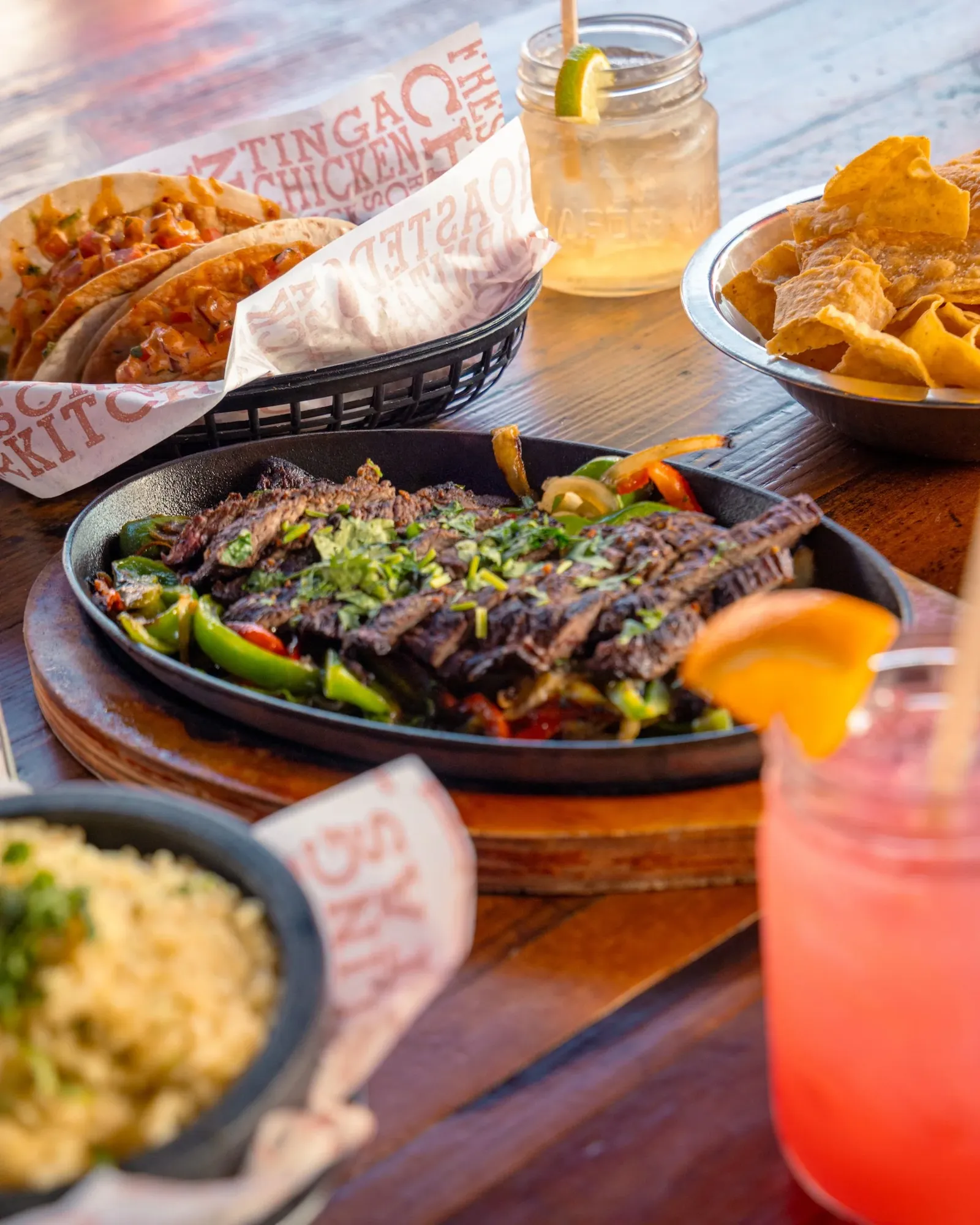 A table with Mexican food including grilled beef with vegetables on a hot skillet, tacos with meat and toppings in a basket lined with paper, a bowl of crispy tortilla chips, and beverages including a