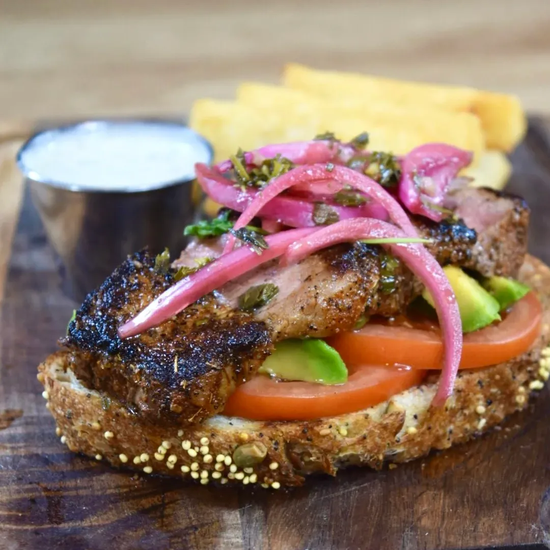 Close-up of a taco with grilled fish, sliced tomatoes, avocado, pickled pink onions, and herbs, served with a side of fries and a small cup of white sauce on a wooden surface.