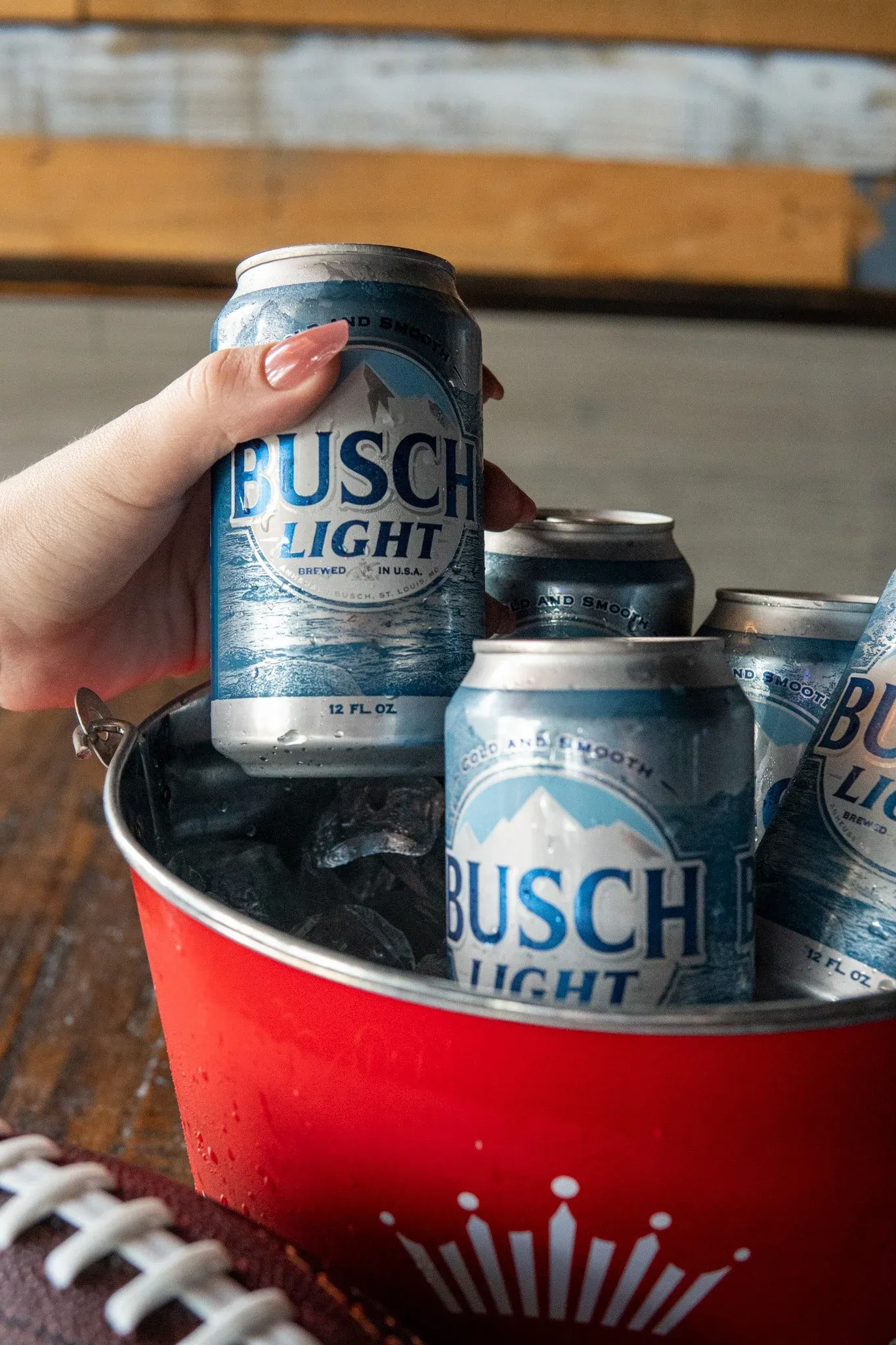 Hand holding a can of Busch Light beer among other cans inside a red bucket.