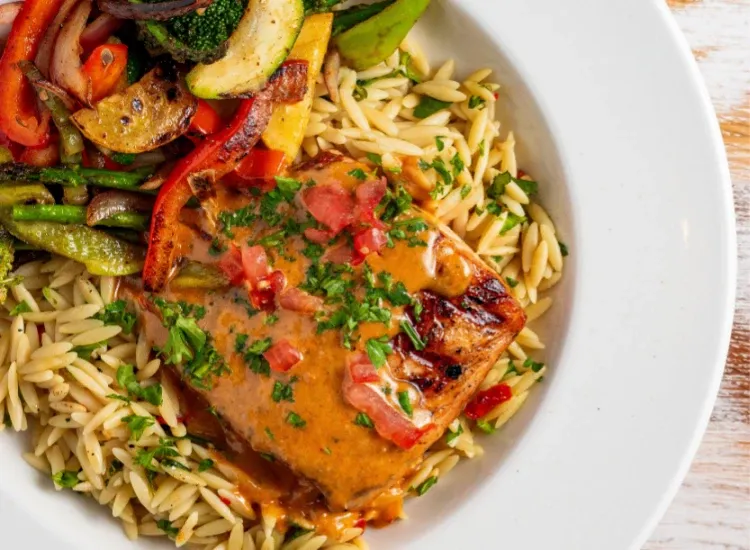 Grilled wild-caught salmon filet topped with a rich sauce, served over orzo pasta and accompanied by grilled vegetables, showcasing Acropolis Taverna's seafood specialties.