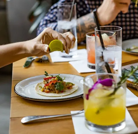 Person squeezing lime over a taco with vegetables and cilantro at a dining table with drinks.