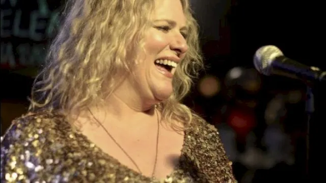 A woman with curly blonde hair laughing on stage, wearing a gold sequined top, with a microphone in front of her at a lively event.