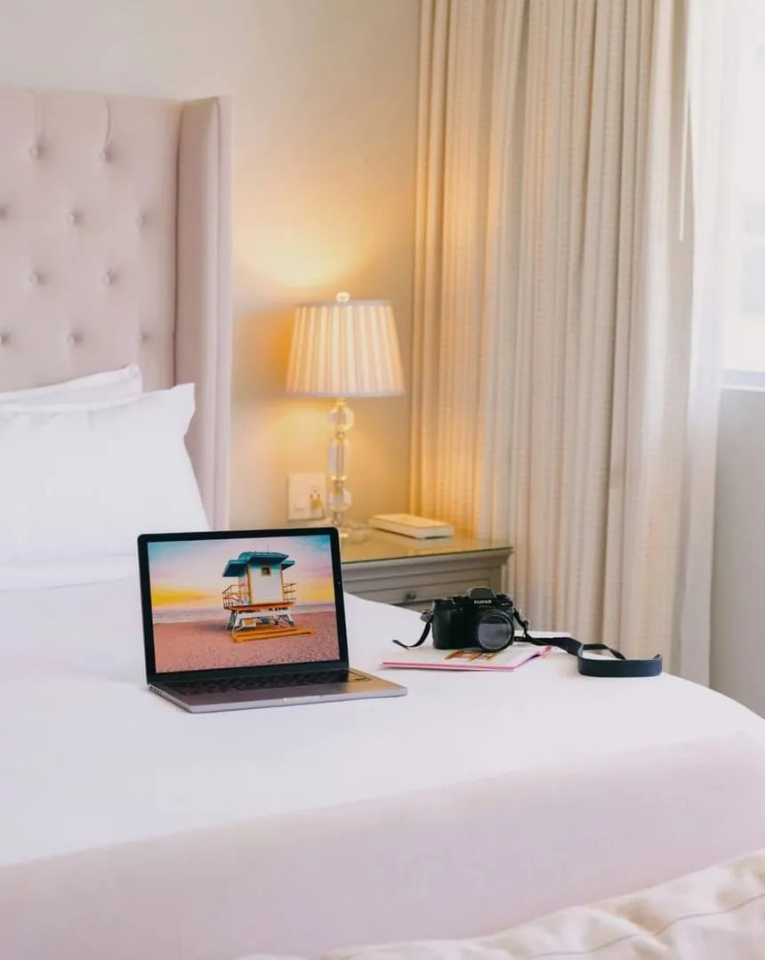 Laptop displaying a beach photo and a camera resting on a plush white bed in a cozy room at The Plymouth Hotel South Beach