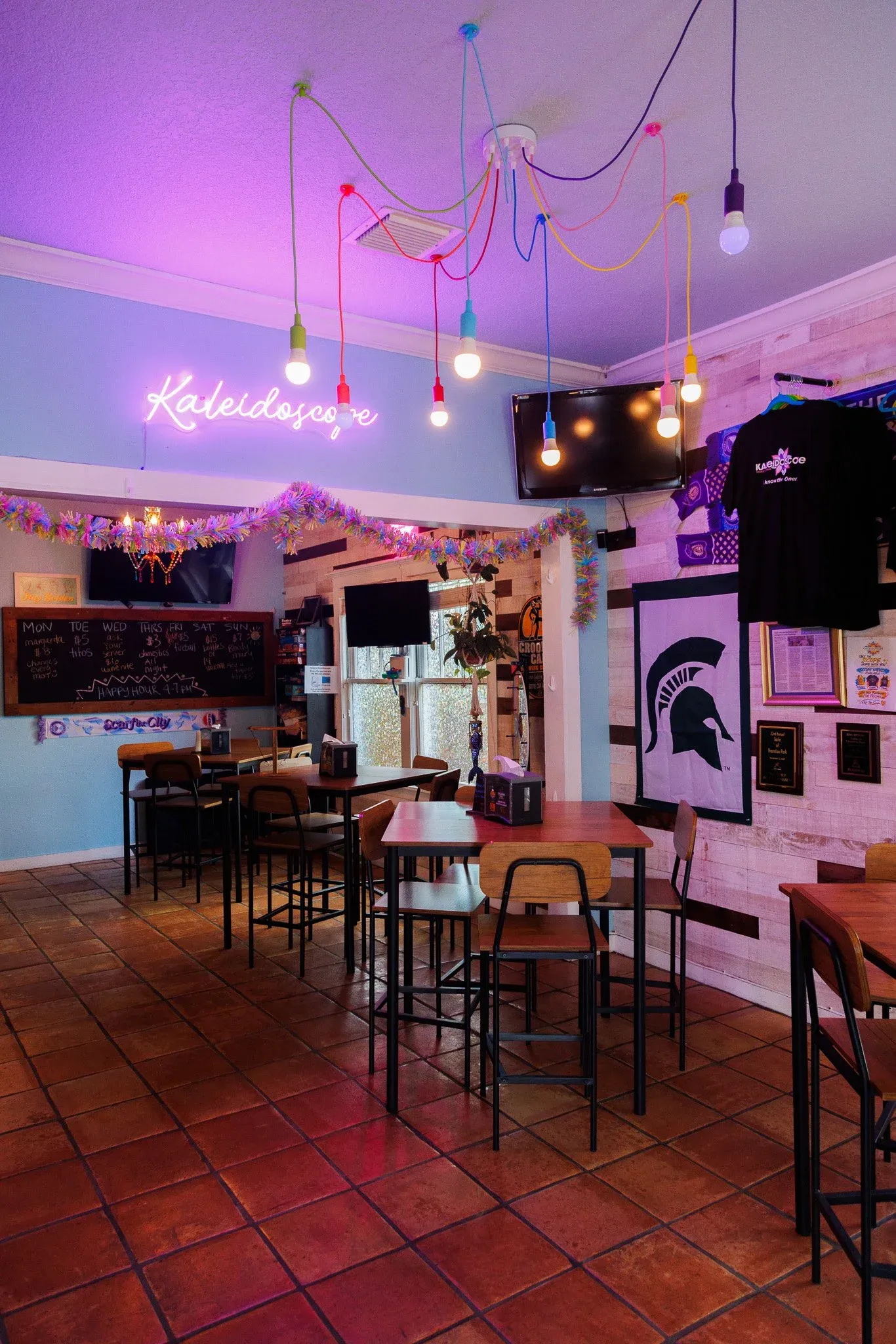 Interior view of Kaleidoscope Thornton Park with wooden tables and chairs, decorated with a purple neon sign that reads 'Kaleidoscope', colorful string lights on the ceiling, a flat-screen TV, and fra