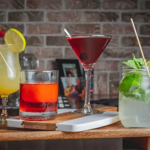A variety of colorful cocktails on a wooden table with a brick wall background, including a lemon-adorned drink, a red drink in a short glass, a dark red martini with a cherry, and a mojito with mint 