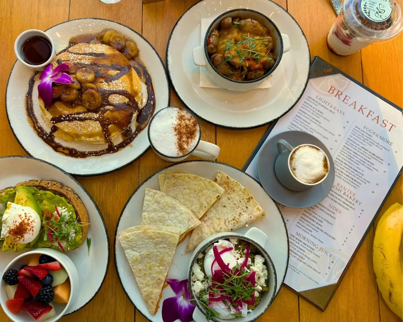 Breakfast spread including pancakes with bananas and syrup, a bowl of soup, a coffee drink, a plate of quesadillas, avocado toast with poached eggs, fresh fruit, a banana, and a glass of layered parfa
