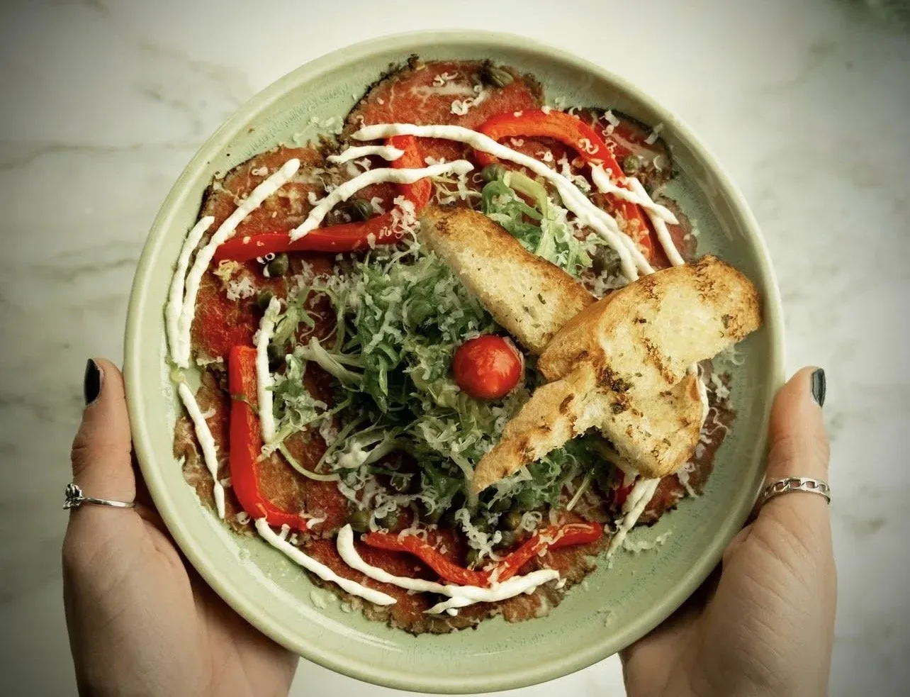 A person holding a bowl of salad with romaine lettuce, red bell peppers, cherry tomato, shredded cheese, sliced cucumbers, and croutons with a drizzle of dressing.