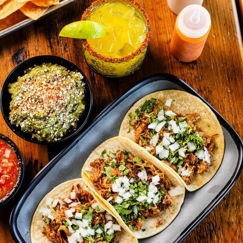 Shredded pork tacos, guacamole, red salsa, and a cocktail, top view.
