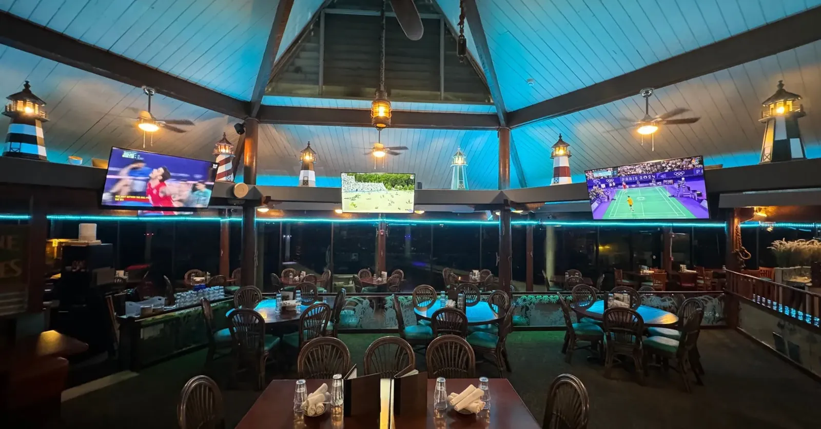 Dimly lit restaurant with a central stage, blue neon accents, and large TVs showing sports.