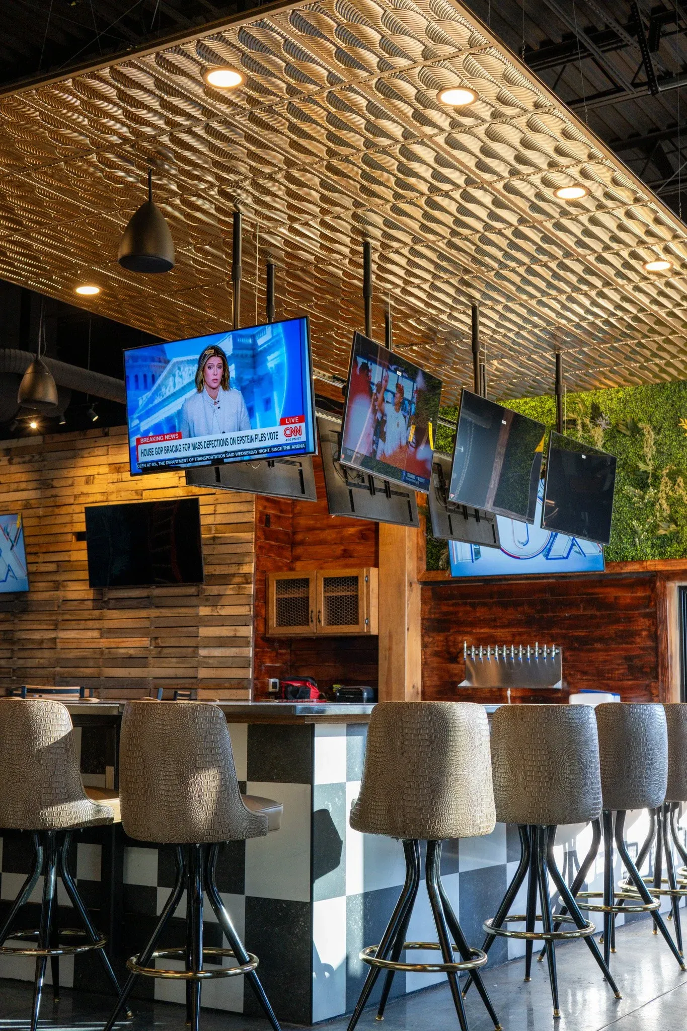 Interior of a modern bar with a row of beige textured bar stools in front of a black and white checkered counter. Multiple mounted flat-screen TVs display news and entertainment, and a decorative wood