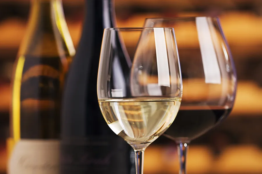 Wineglasses and wine bottles on display for serving a wine tasting. Red and white fermented grape beverages are poured into two sparkling glasses. Warm, orange brown wooden cellar wine racks are refle