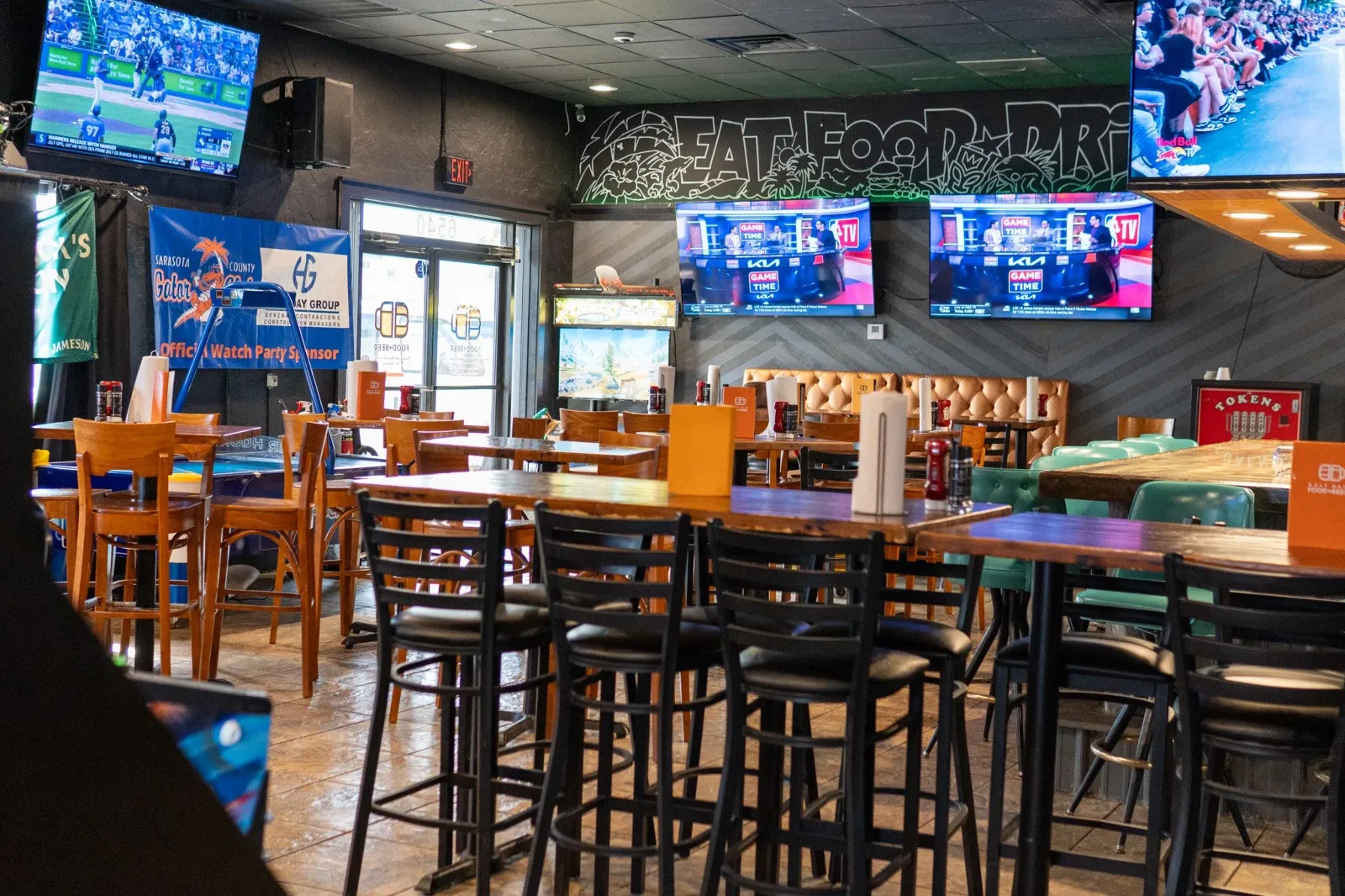 An empty sports bar with multiple flat-screen TVs showing baseball and football games. There are wooden and metal barstools and tables, a dart machine, and a neon sign that reads 'Eat Food.' The bar h