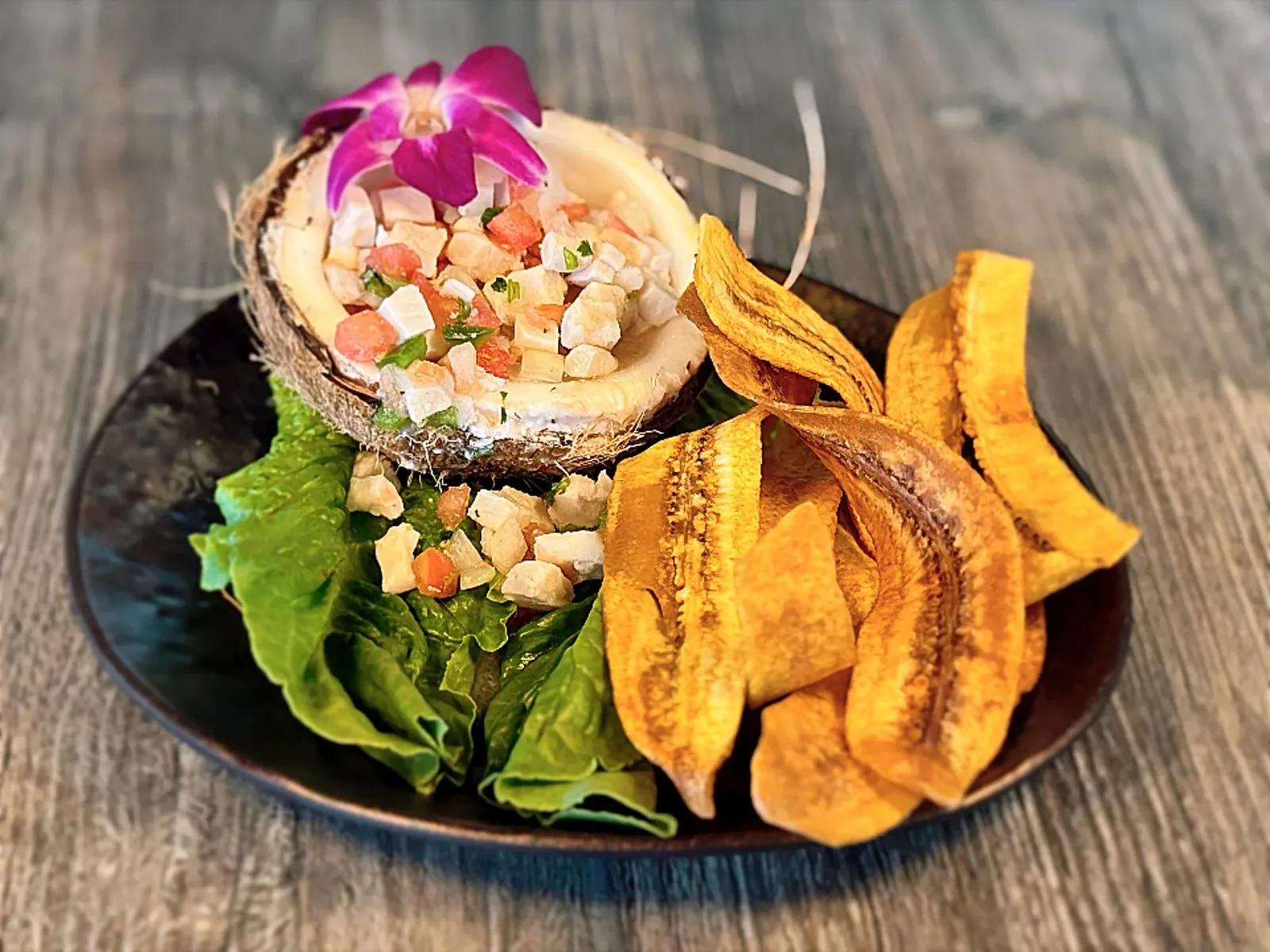 Ceviche served in a coconut shell on a leafy bed, garnished with a purple orchid flower, accompanied by crispy plantain chips on a black plate.