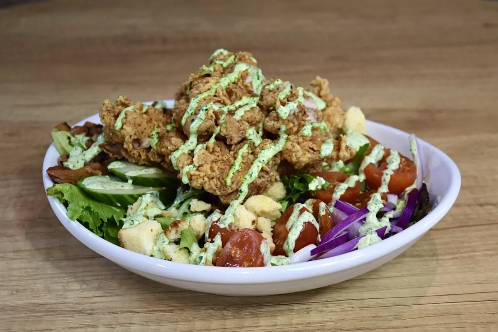 A salad with mixed greens, cherry tomatoes, shredded purple cabbage, cucumber slices, croutons, and fried chicken strips drizzled with green dressing in a white bowl on a wooden surface.