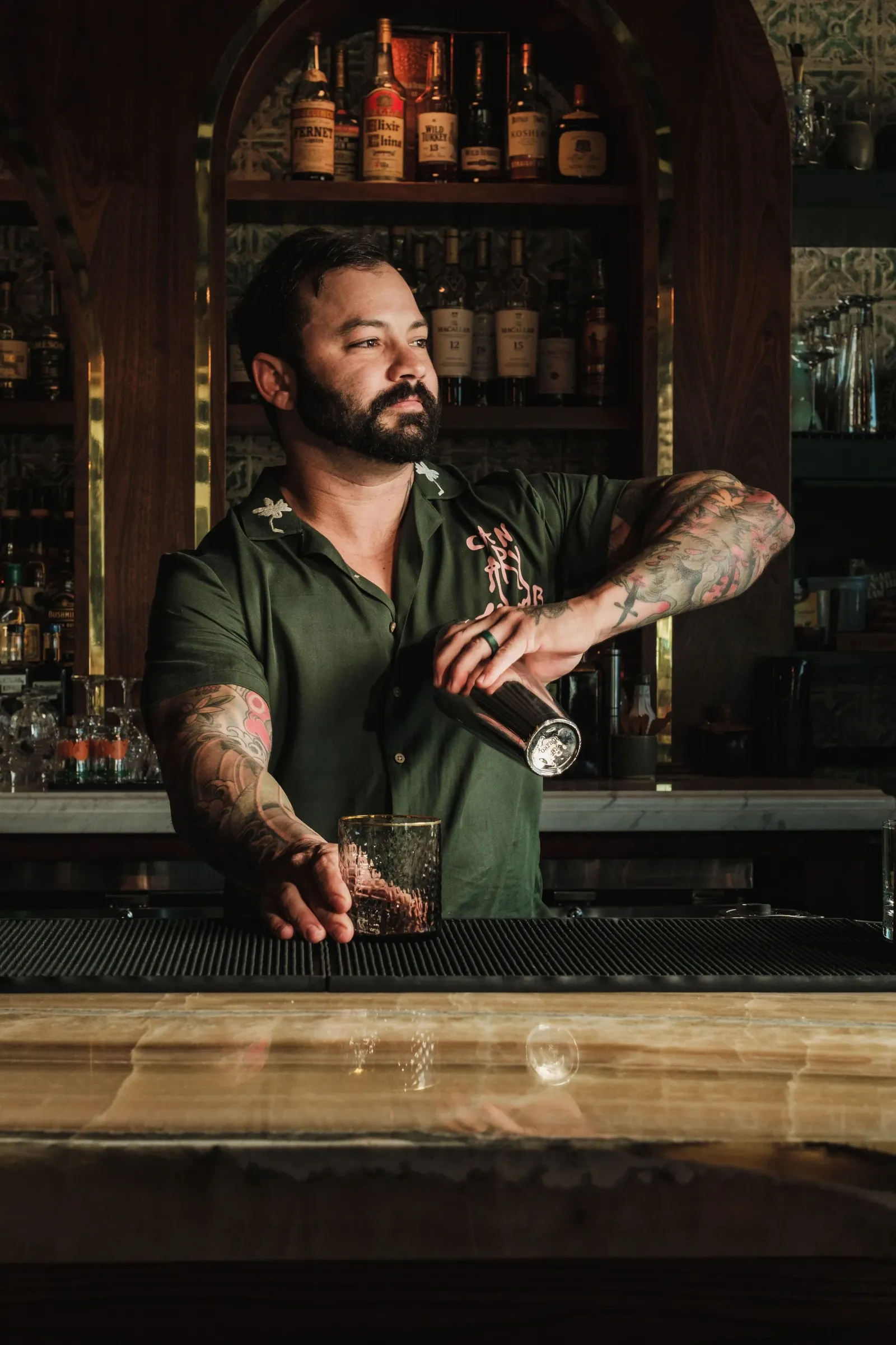 A man with tattoos on his arms and a beard, wearing a dark green shirt with decorative embroidery, stands behind a bar holding a glass of dark-colored drink and an ice scoop. The bar has a wooden back