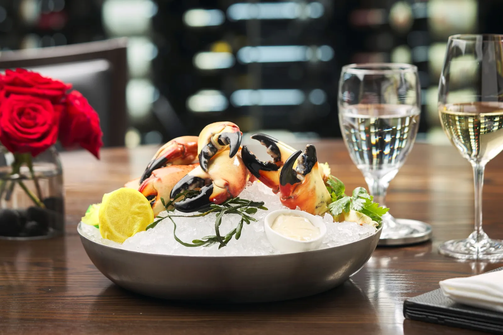 a platter of fresh Florida stone crab claws on a bed of crushed ice with fresh seaweed, a wrapped lemon and a small ramekin of mustard sauce sitting on a table with a glass of wine and fresh red roses