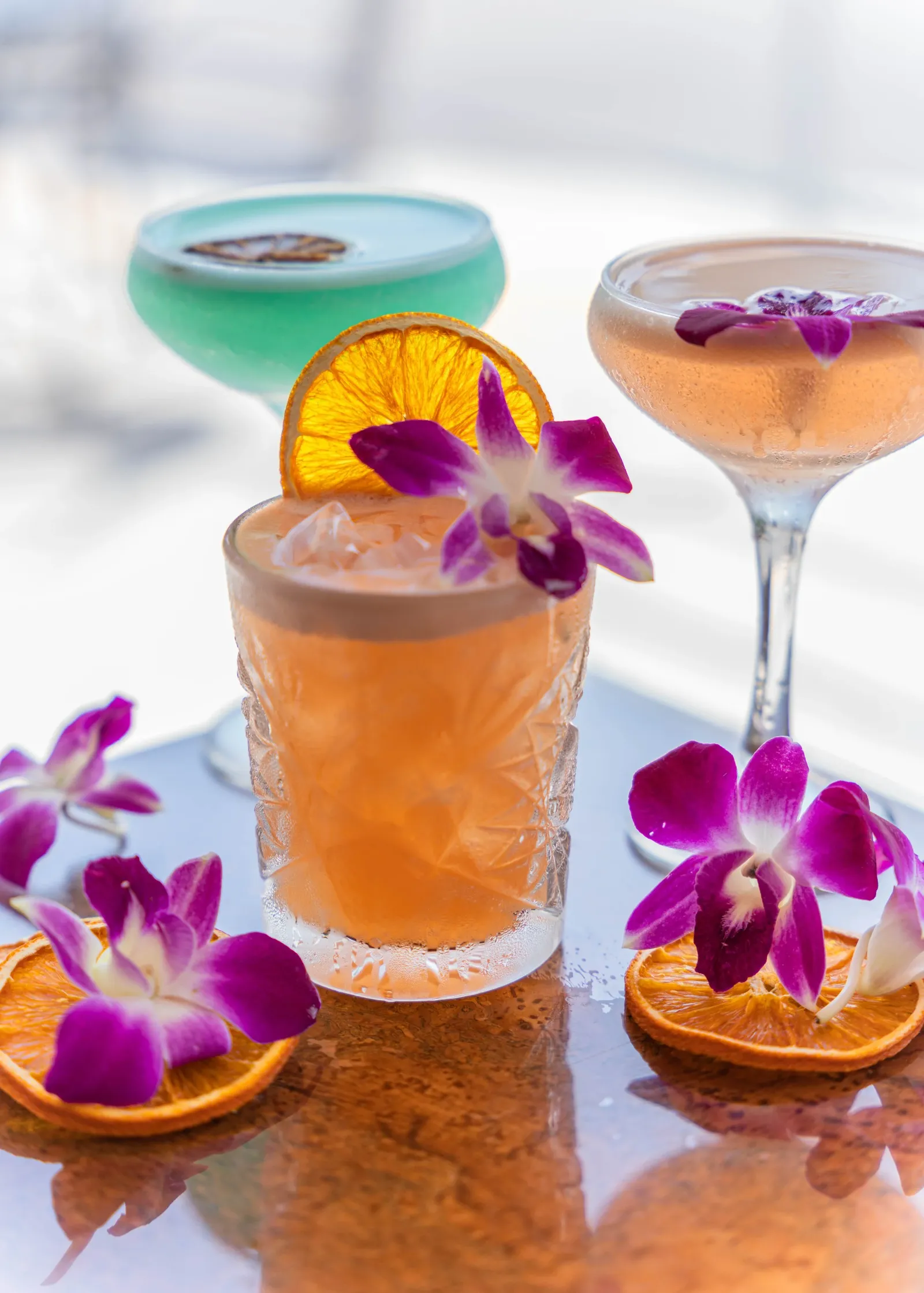 Three colorful cocktails garnished with dried orange slices and purple orchids, with more orchids and orange slices on a reflective surface.