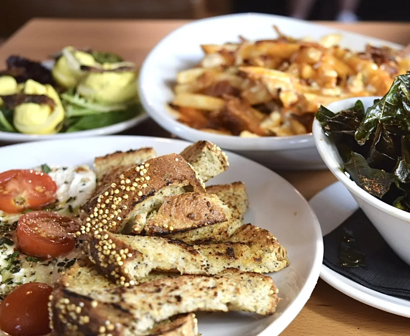 Assorted dishes including grilled chicken with seasoning, cherry tomato salad, roasted potato dish, sautéed greens, and other vegetables on white plates and bowls