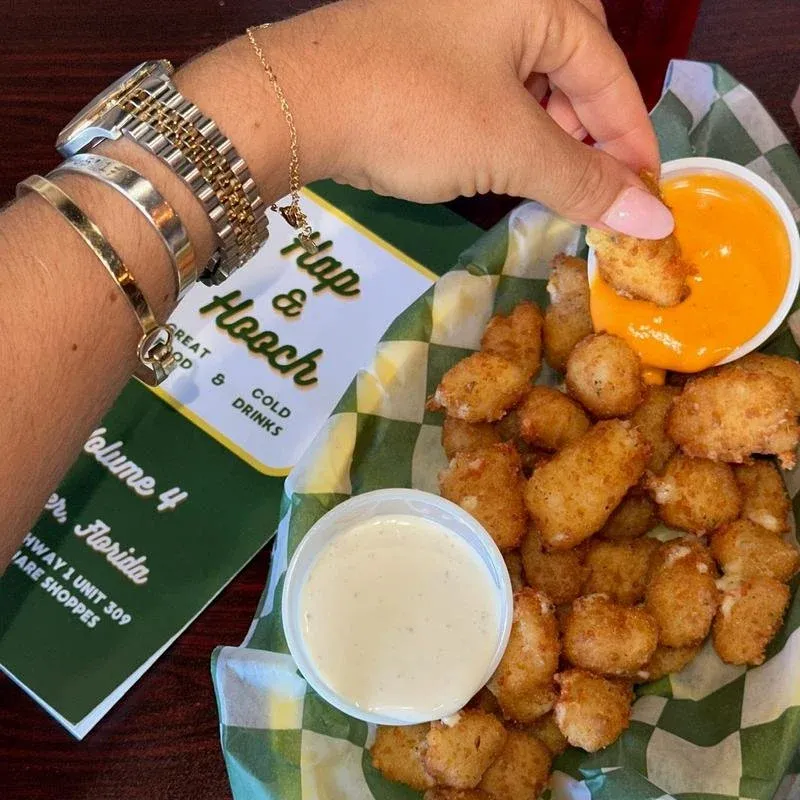 A hand holding a piece of fried food dipped in orange cheese sauce with a basket of fried nuggets and two dipping sauces, one creamy and the other with orange cheese, on a paper-lined basket. A menu o