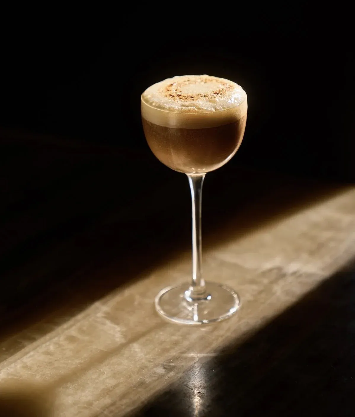 A cocktail in a coupe glass with a frothy top, garnished with a sprinkle of spices or cocoa powder, against a dark background with a wooden surface.