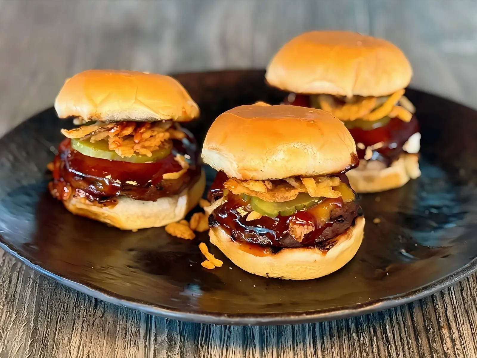 Three beef sliders on a dark plate with pickles and crispy onions.