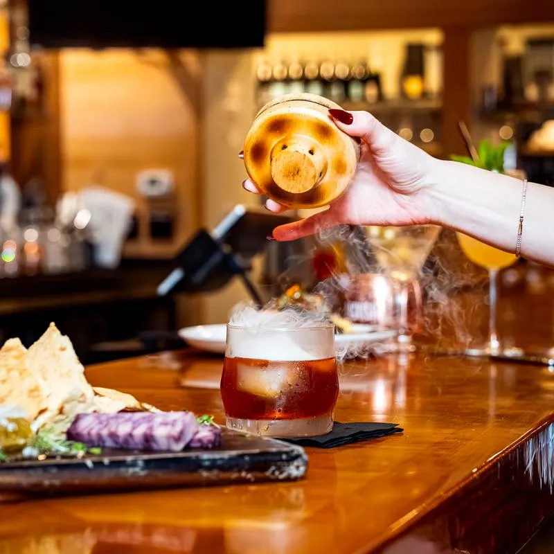 A hand lifting a smoked cocktail lid on the bar.