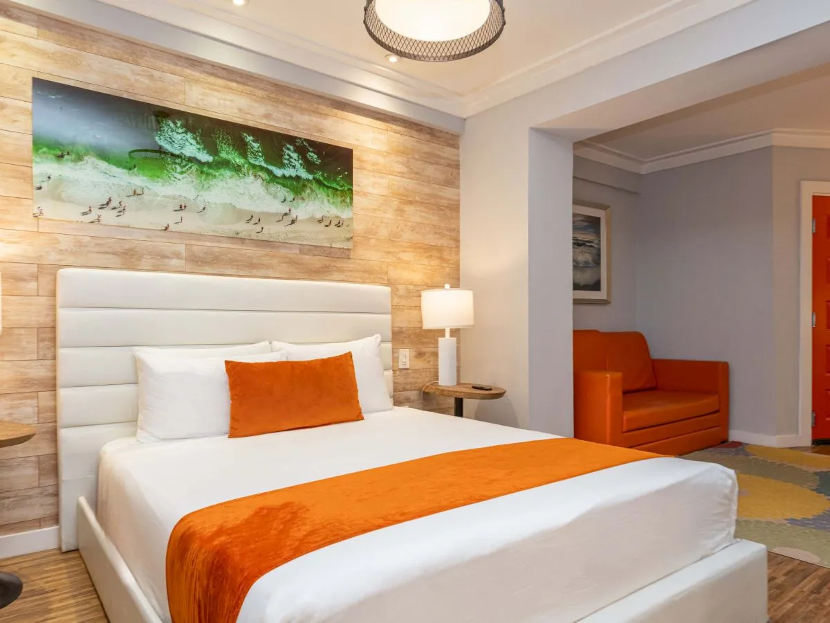 A modern hotel room with a white bed, orange accents, wall lamps, and a seating area near the doorway. 140 characters.
